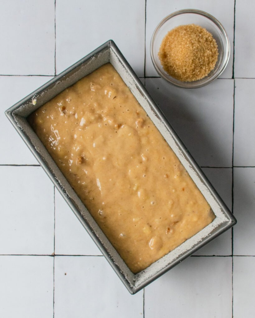 Batter in a loaf pan with turbinado sugar in a small bowl next to it.