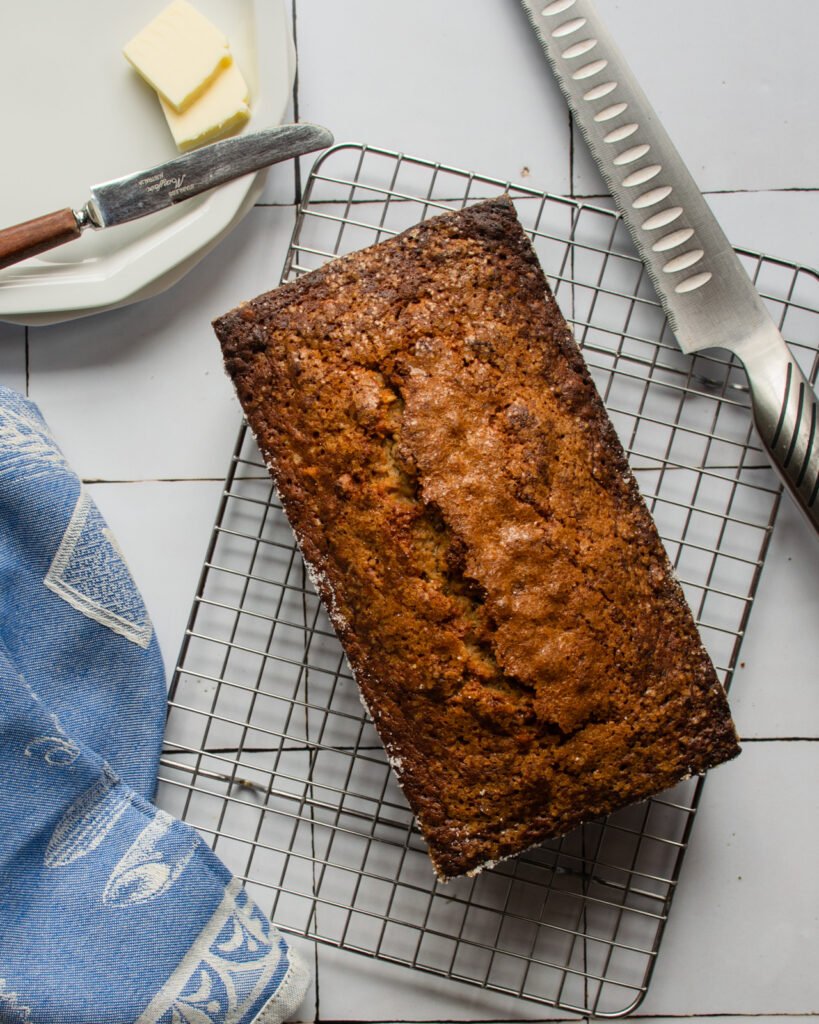 Banana bread on a cooling rack with blue towel, bread knife, small white plate with butter.
