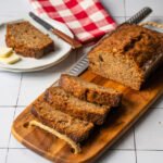 A loaf of banana bread on a wooden cutting board with a few slices cut, along with a small white plate holding one piece of banana bread.