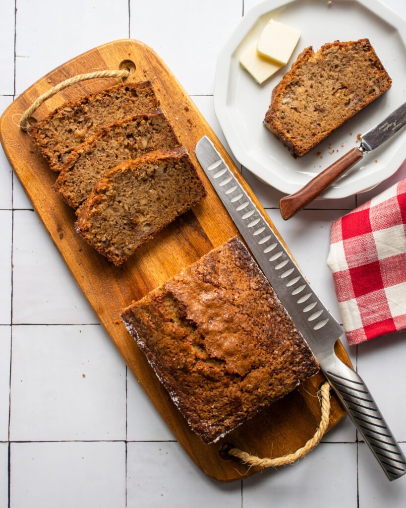 Banana bread on a cutting board with a few slices already cut and one on a white plate with butter.