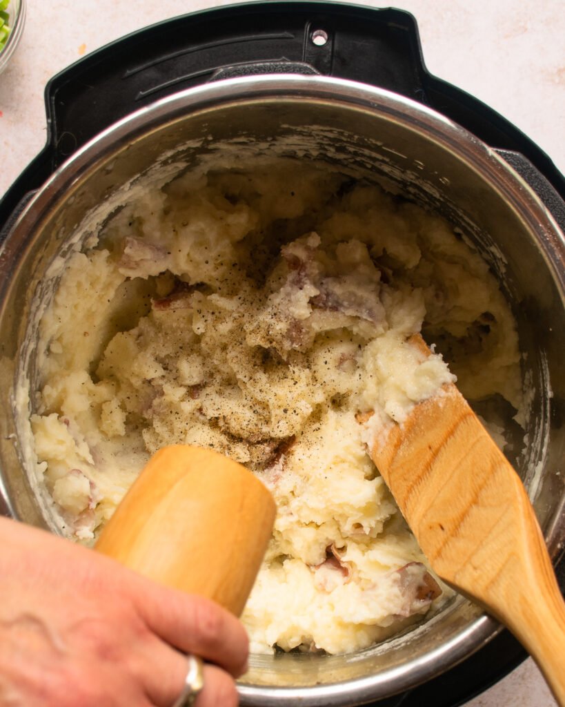 A wooden spoon sticking out of a pressure cooker while a hand grinds fresh black pepper into the pot.