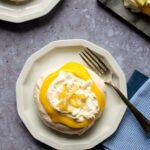 Pavlova with lemon curd and whipped cream on a white plate on a table with blue napkins and a fork.