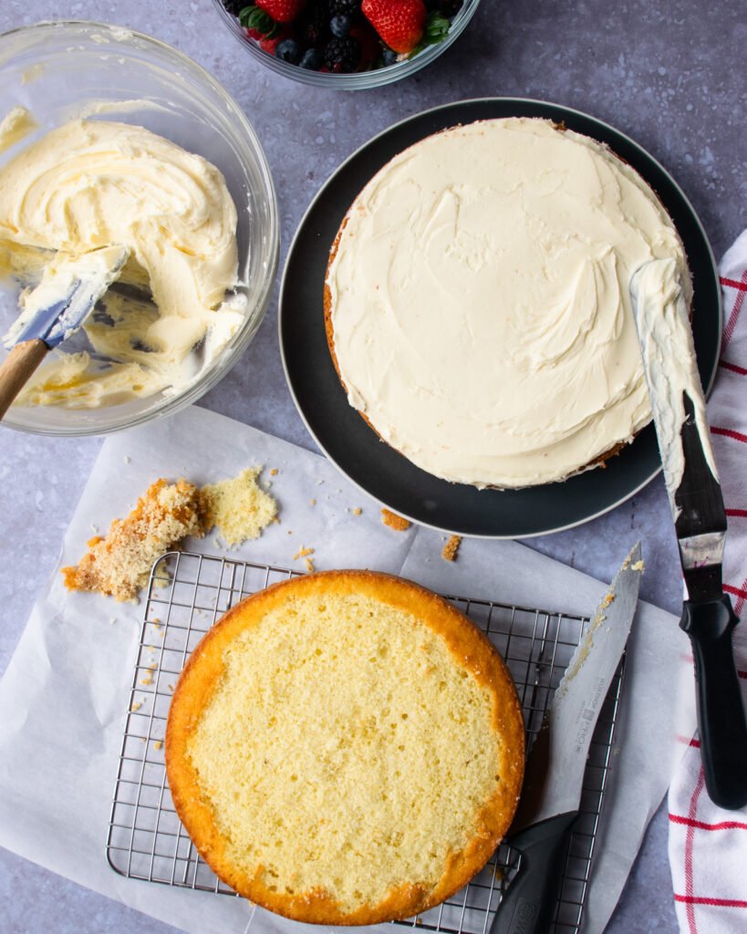 A layer cake being assembled with buttercream frosting on one layer and another layer of cake ready to be placed on top.