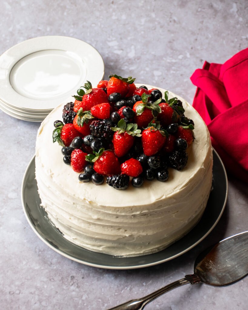 A vanilla Layer cake with berries piled on top and a red napkin beside it.