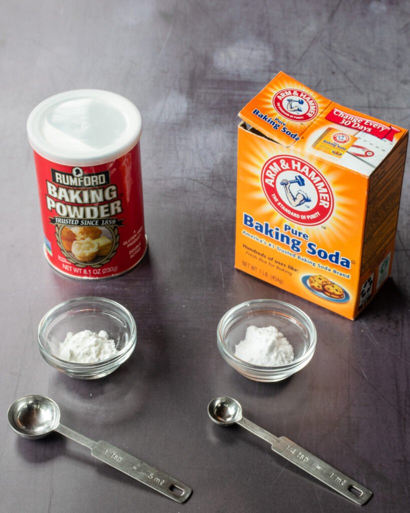 A can of baking powder and a box of baking soda with little dishes of each in front of them with measuring spoons..