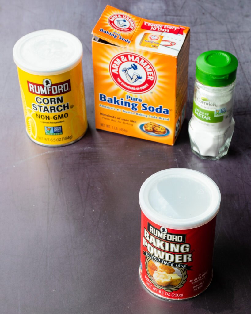 A can of baking powder in front of a can of cornstarch, a box of baking soda and a bottle of cream of tartar.