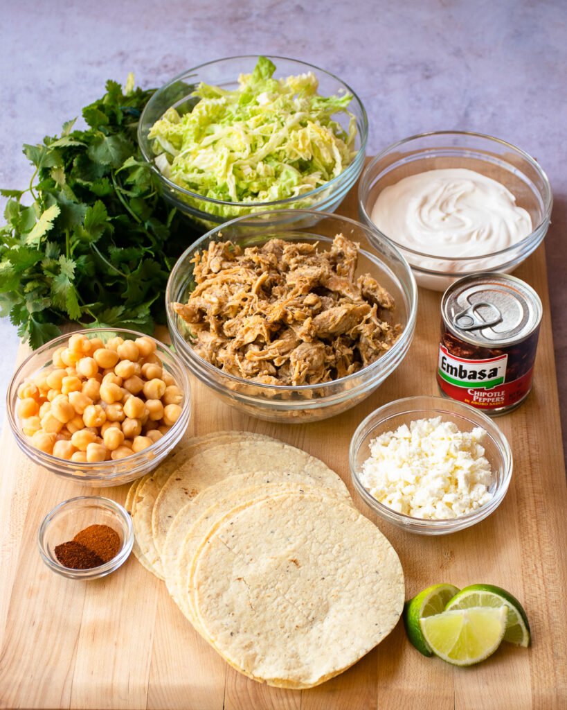 Ingredients for tostadas on a board - tortillas, chickpeas, shredded chicken, cabbage, cheese, chipotle pepper, lime, sour cream and cilantro.