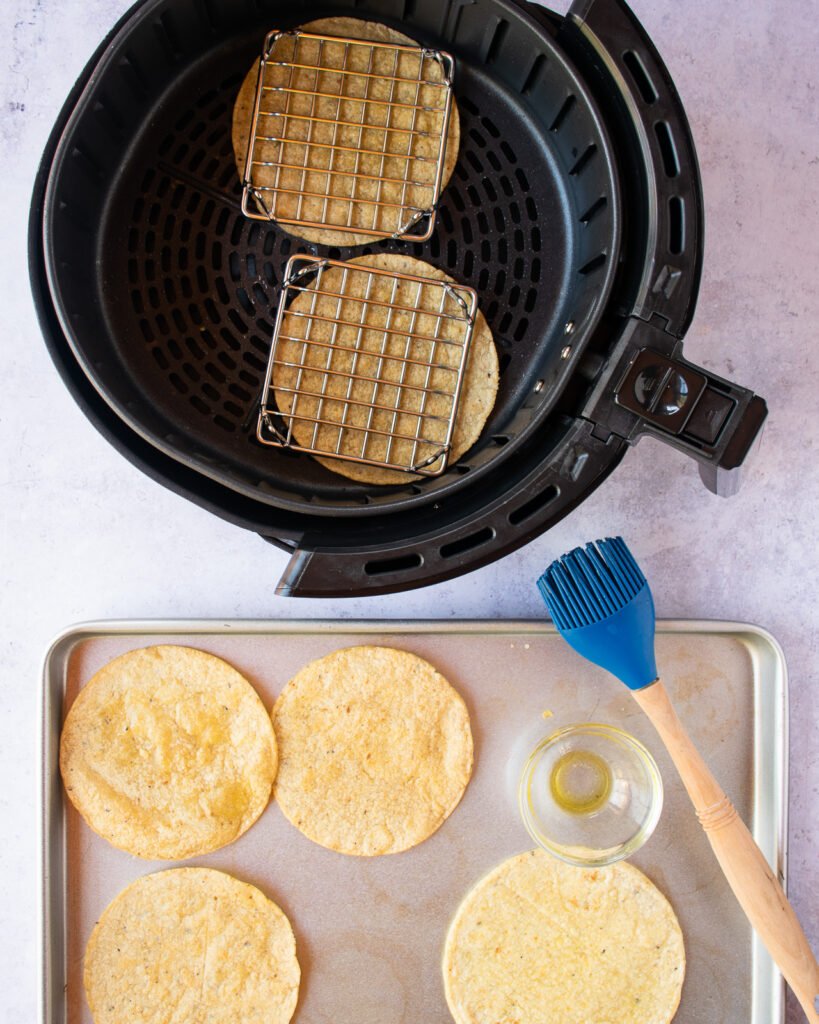 Two tortillas in an air fryer with more tortillas on a sheet pan with olive oil and a basting brush near by.