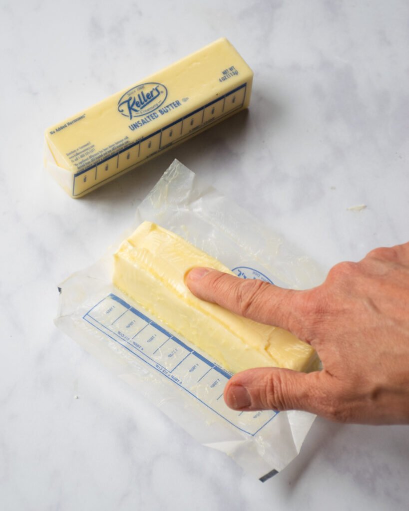 A finger pressing a stick of butter on a counter.