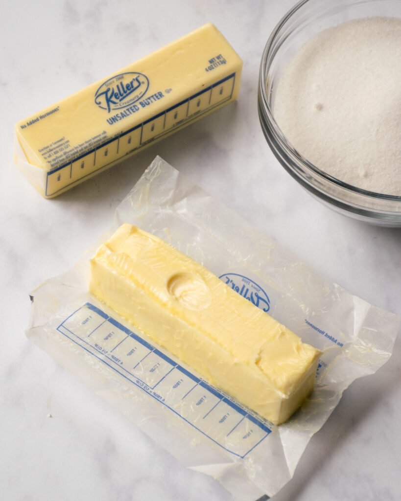 A stick of butter with a finger indentation in it. Another stick of butter and a bowl of sugar next to it.