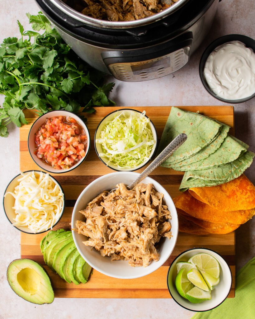 All the ingredients for instant chili lime chicken tacos on a wooden cutting board next to a pressure cooker.