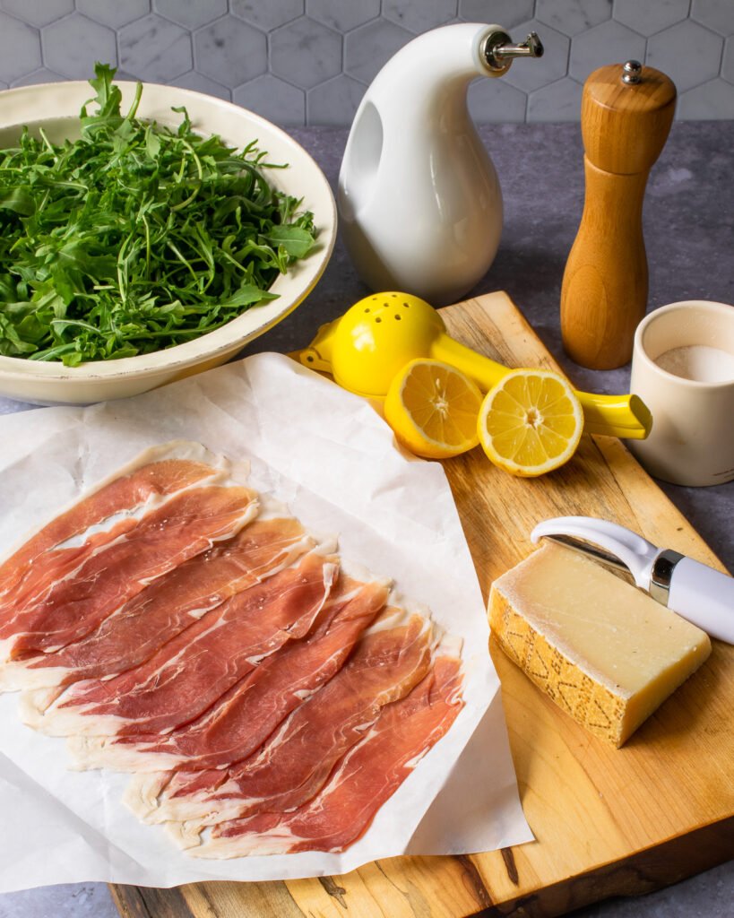 Ingredients on a cutting board - prosciutto, parmesan cheese, lemon halves - a bowl of arugula, olive oil, salt and pepper near by.