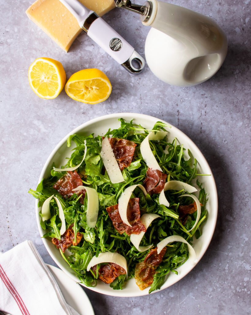 A white plate with arugula parmesan prosciutto salad on a grey counter.