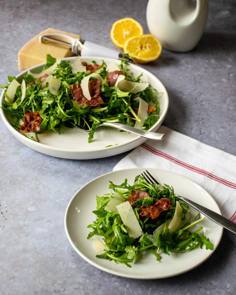 A white salad plate with arugula parmesan prosciutto salad in the foreground and a larger serving plate of salad behind it.