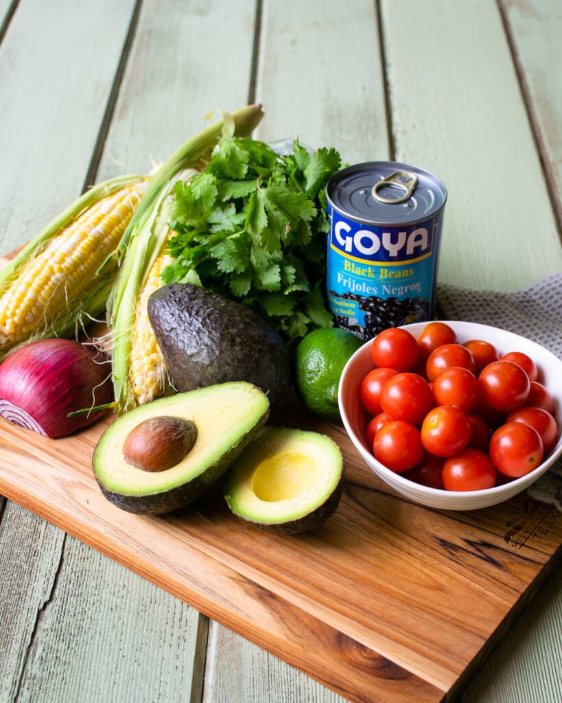 Avocado, tomatoes, ears of corn, cilantro, a red onion, a can of black beans and a lime on a wooden cutting board on a green wooden table.