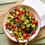 Avocado Tomato salad in a terra cotta rimmed white bowl on a placemat on a green wooden table.