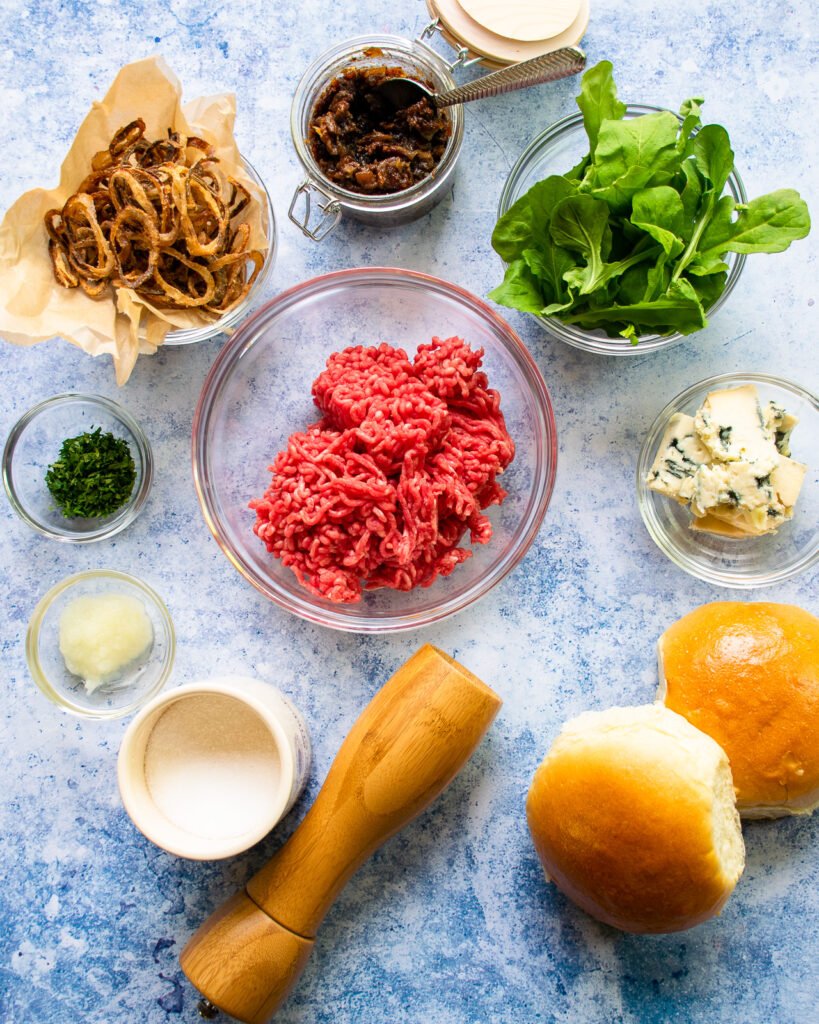 Ingredients on a blue counter - ground beef, salt, pepper, brioche buns, blue cheese, arugula, bacon jam, crispy shallots, parsley and grated onion.