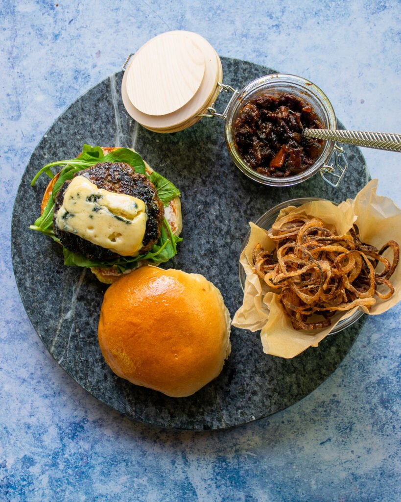 A bacon blue burger being assembled - burger on bun with a jar of bacon jam and a bowl of crispy shallots.