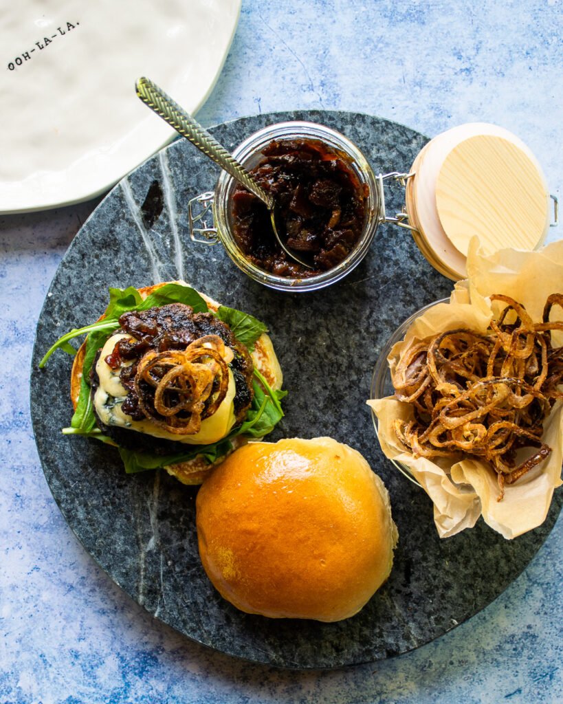 Looking down on a burger with bacon jam on it next to a jar of bacon jam and some crispy shallots.