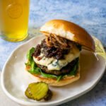 Bacon Blue Burger on an oval white plate with pickles on the side and a beer in a glass.