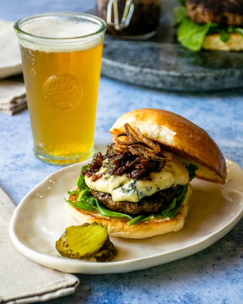 A bacon blue burger with a side of pickles and a glass of beer.