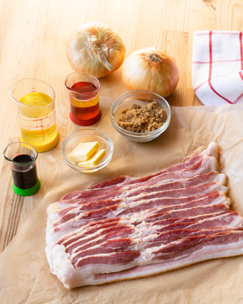 A pound of bacon, balsamic vinegar, water, brown sugar, onion, butter and cider vinegar on a wooden counter with a red and white kitchen towel.