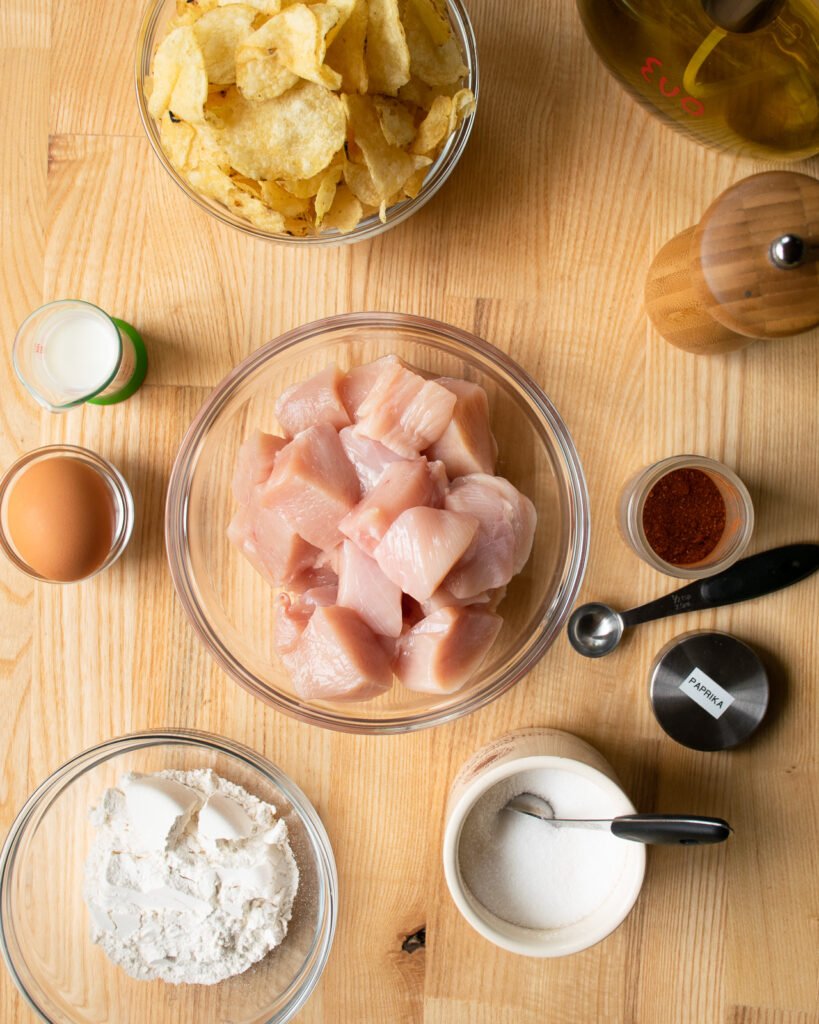 Ingredients in bowls on a butcher block countertop - chicken, egg, potato chips, paprika, salt, pepper, flour and milk.