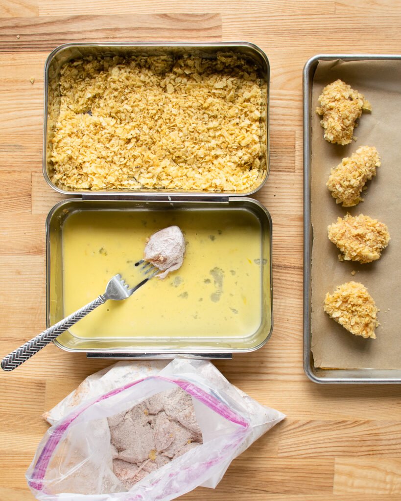 A dredging station on a countertop - floured chicken pieces in a bag, a shallow dish of egg wash and a shallow dish of potato chip crumbs - next to a sheet pan of coated chicken nuggets.