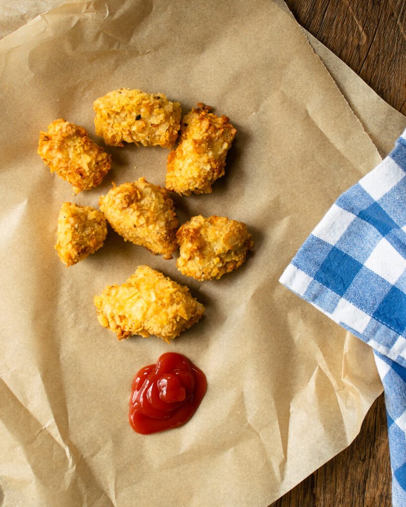 Chip crusted chicken nuggets on brown parchment paper with ketchup and a blue checkered napkin.