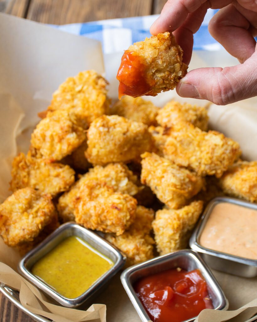 A hand holding up a chicken nugget over a basket of nuggets with dipping sauces.