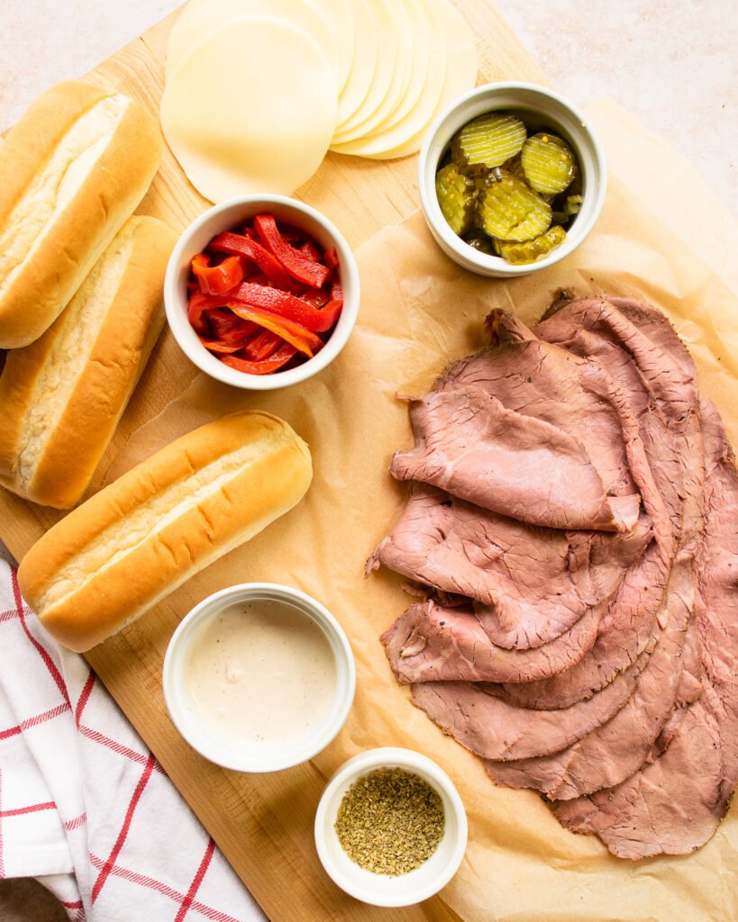 Roast beef, red pepper, pickle chips, horseradish mayonnaise, oregano and steak rolls on a cutting board with a red windowpane towel nearby.