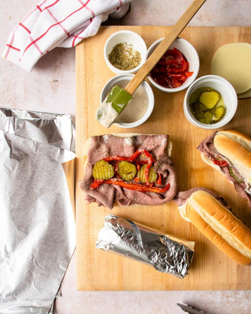 One roast beef sandwich being assembled on a cutting board next to a second sandwich wrapped in foil.