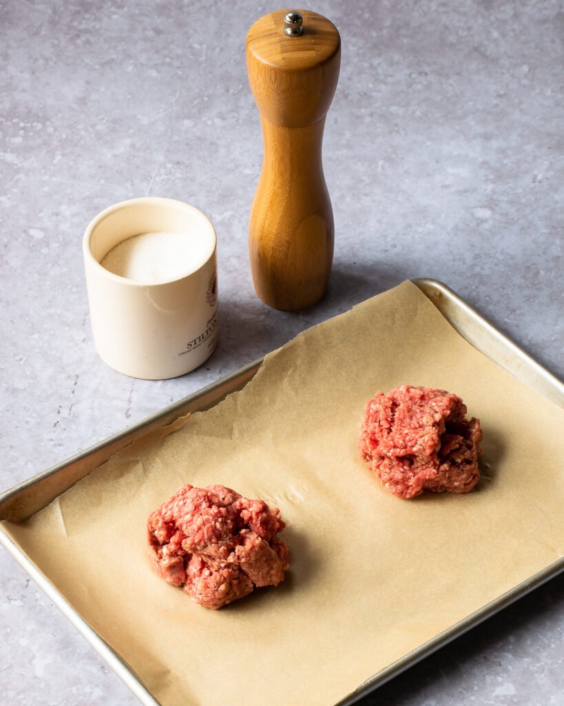Two lumps of ground beef on a parchment paper-lined baking sheet with salt and pepper next to it.