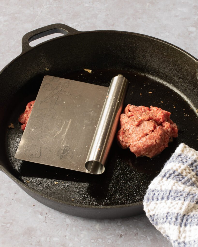 Two smash burgers being pressed in a black cast iron pan.