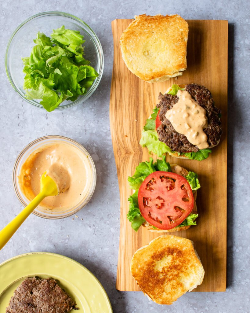 Building a smash burger - a wooden board with two hamburger set ups next to a bowl of burger sauce and a bowl of shredded lettuce.