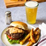 A smashburger with French fries on a white plate with a salt shaker and a glass of beer nearby.