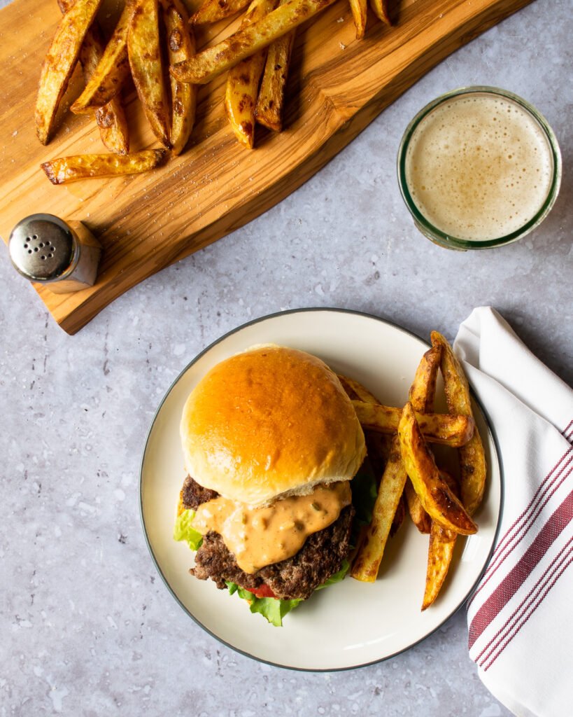 A smash burger with fries on a white plate with a glass of beef and more french fries on a wooden board.