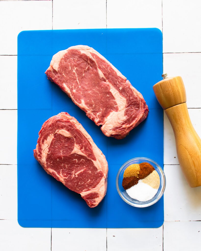Two ribeye steaks on a blue cutting board with a bowl of spices and a pepper mill.