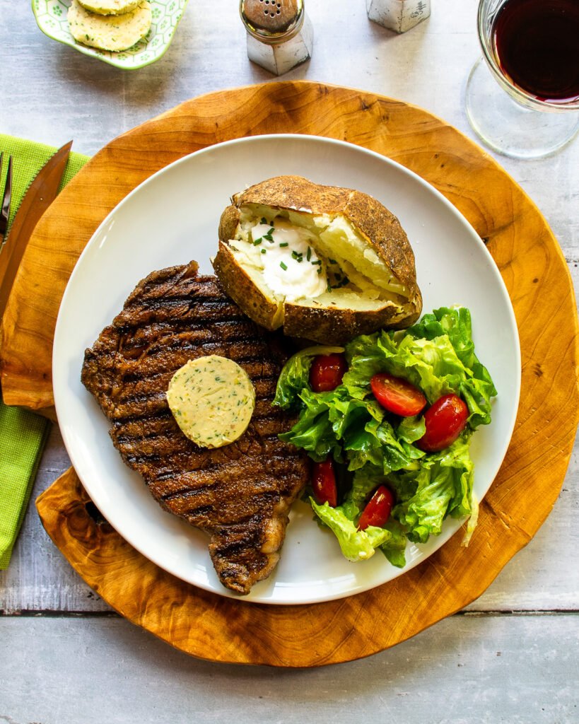 A dinner plate with a baked potato, salad and a ribeye steak with compound butter on top.