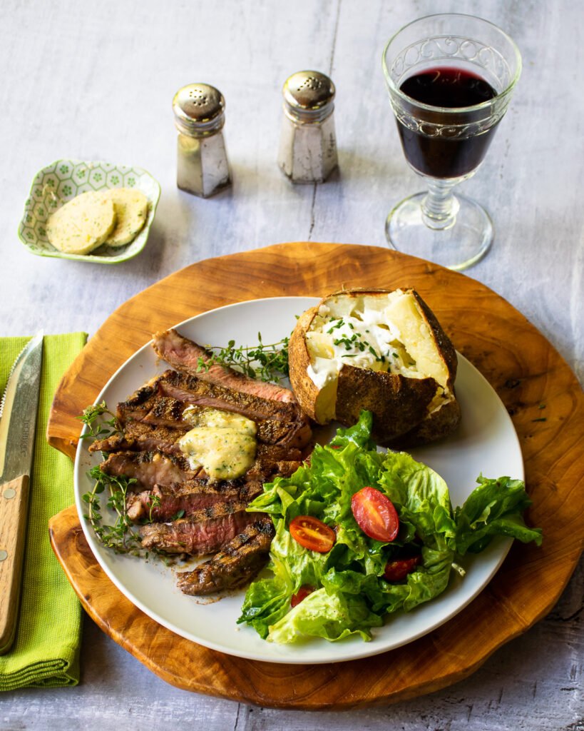 A dinner plate with sliced smoke spiced grilled ribeye, a baked potato, salad, glass of wine, salt and pepper shakers and a dish of compound butter.