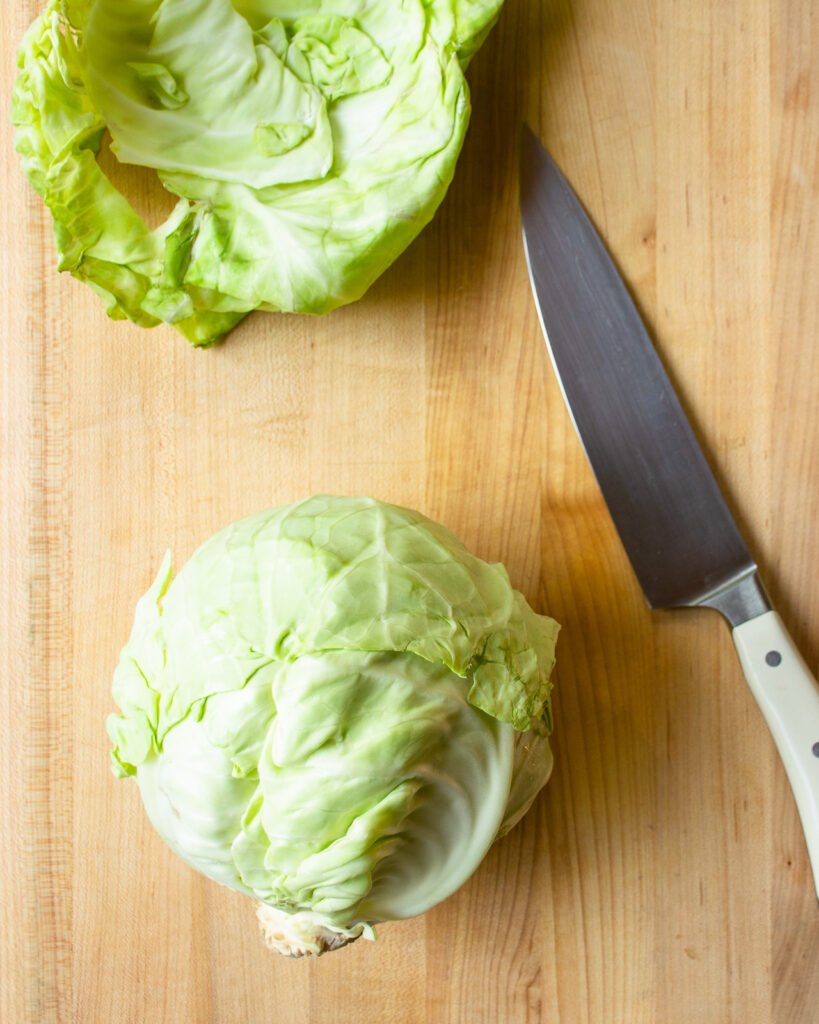 A head of green cabbage next to a knife with the outer leaves removed.