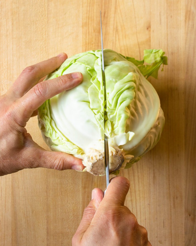 Two hands cutting a head of green cabbage in half.