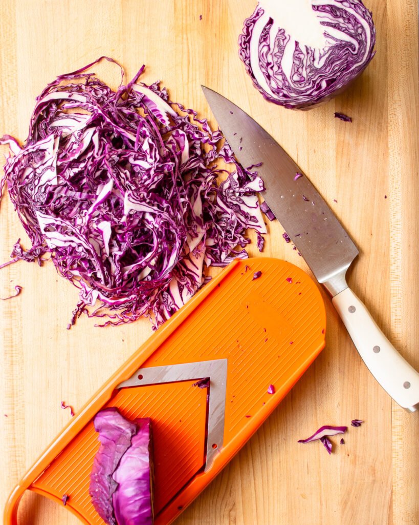 Shredded red cabbage on a butcherblock counter next to a mandolin and a chef's knife.