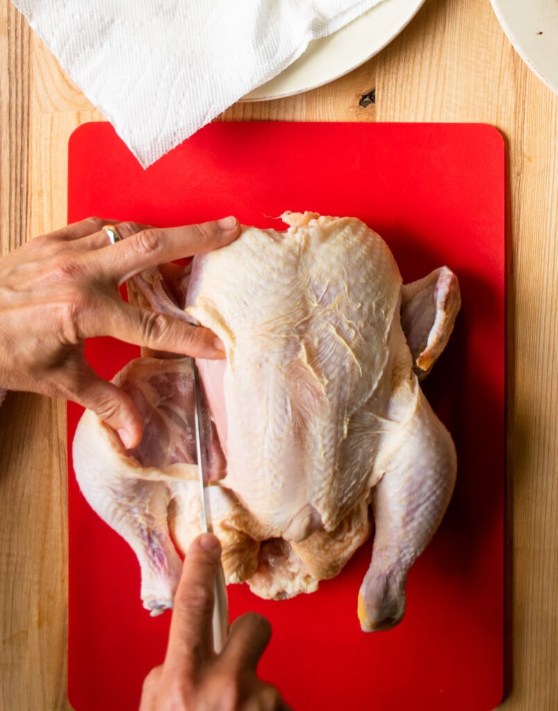 A hand cutting the thigh away from a raw chicken on a red cutting board, the first step in how to cut up a whole chicken.. 