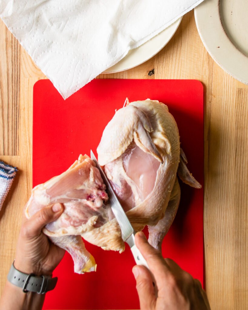 Two hands cutting the leg off a chicken on a red cutting board.