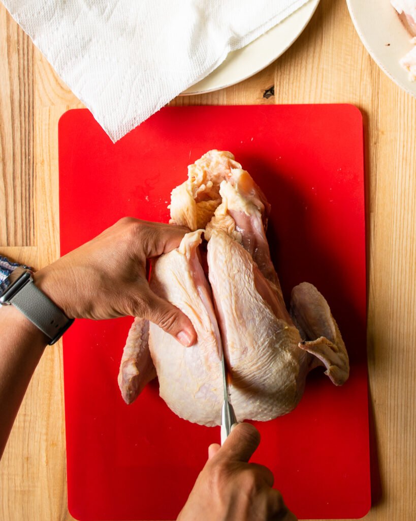 A knife cutting a raw chicken in half along the breastbone on a red cutting board, the third step in how to cut up a whole chicken..