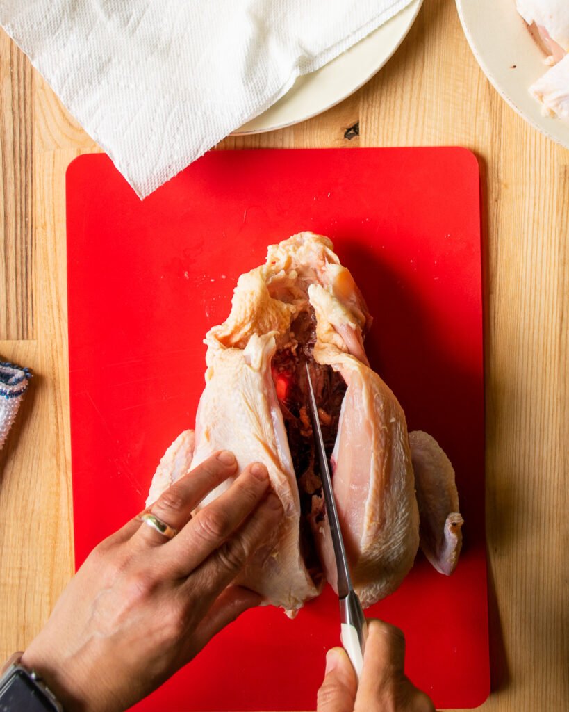 Two hands slicing through the back of a chicken to remove a split breast on a red cutting board.