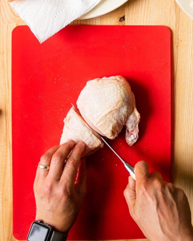 Two hands slicing a chicken breast in half on a red cutting board, the fourth step in how to cup up a whole chicken.