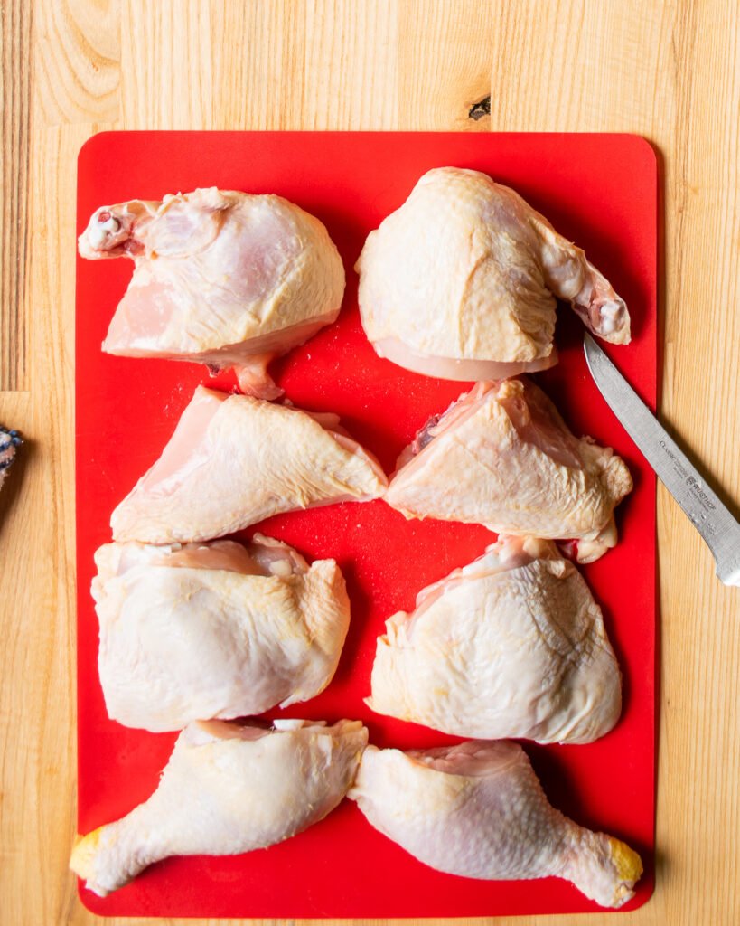 All the pieces of a whole chicken cut into 8 pieces on a red cutting board.