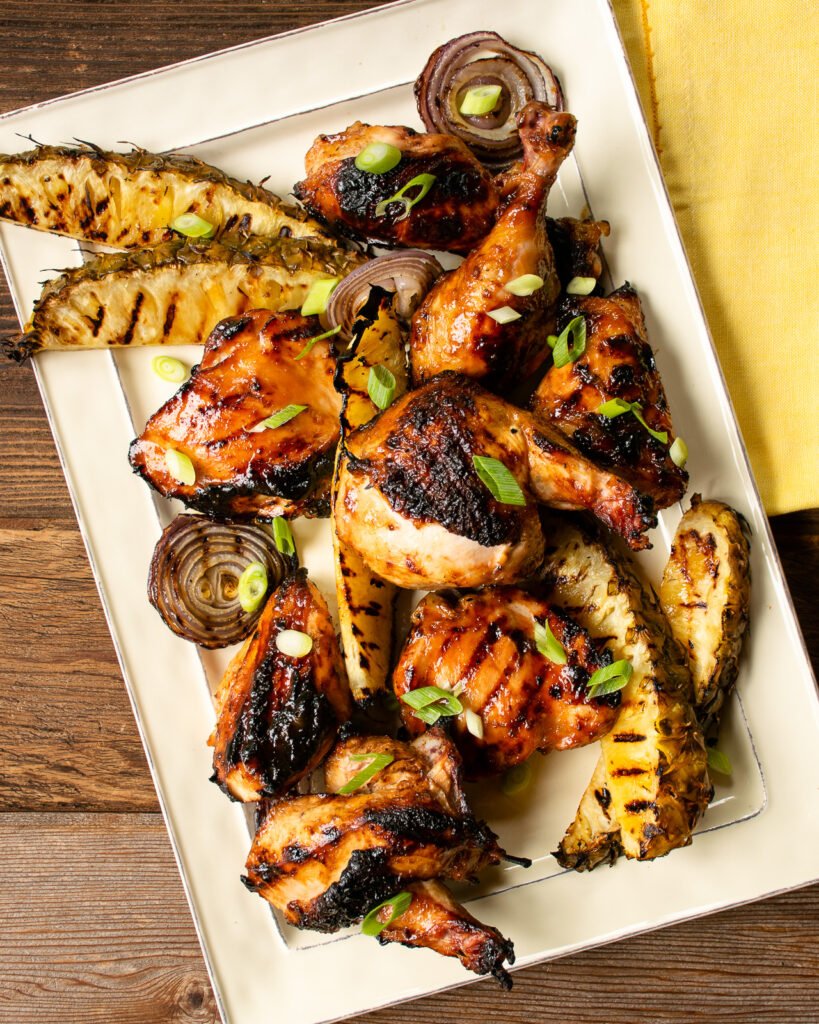 Huli Huli Chicken on a rectangular platter with grilled pineapple and red onion and a yellow napkin.